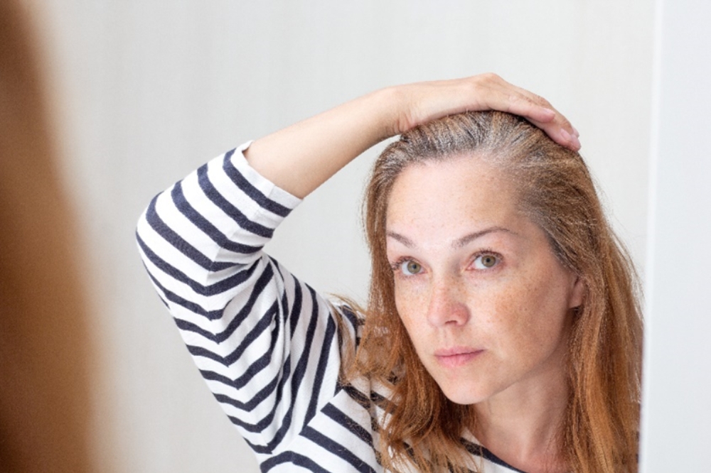 Žena v menopauze si obzerá šediny v zrkadle a zisťuje, že má zhoršenú kvalitu vlasov.