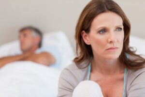 Žena v menopauze sedí na posteli a vyzerá smutne, pretože v partnerskom vzťahu riešia otázku straty libida.