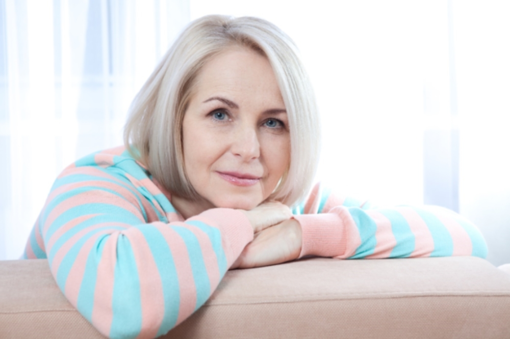 Staršia žena v menopauze sa opiera o rukami o gauč a pozerá sa dopredu s úsmevom na perách, pretože ju netrápi strata libida a iné príznaky menopauzy.