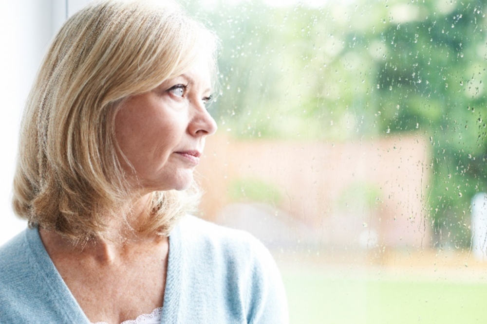 Staršia žena v menopauze pozerá z okna a premýšľa nad obdobím menopauzy a jej príznakmi.